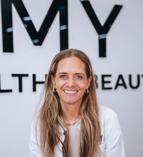 Blonde woman in a white lab coat smiles, standing before a "MY HEALTH BEAUTY" sign.