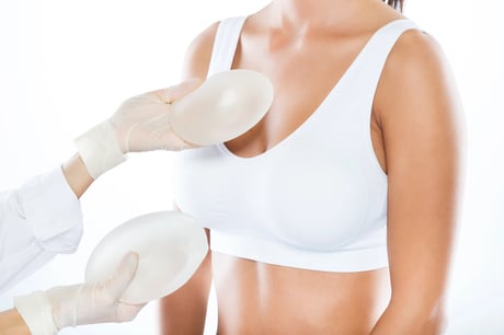 Gloved hands presenting breast implants to a woman in a white sports bra.